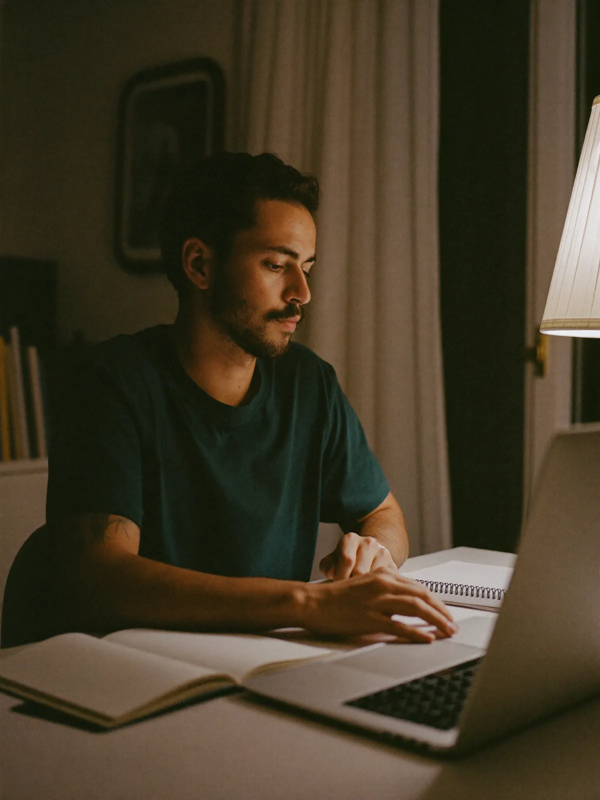 Homem de uns 35 anos estuda à noite no notebook, com um caderno aberto na mesa da cozinha